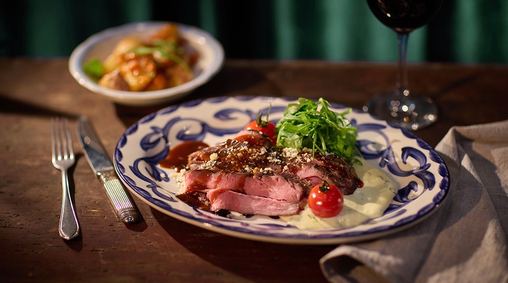 Mahlane steik garneering rukola ja kirsstomatitega sini-valge mustriga taldrikul. Taustal teine roog ja veiniklaas.