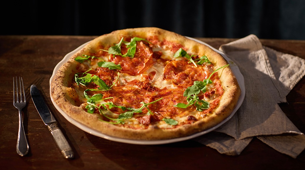 Salaamiviilude ja värske rukolaga pitsa puuservaga taldrikul, kõrval kahvel ja nuga.