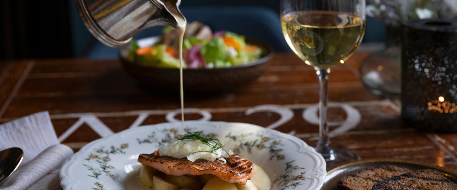 Sauce being poured over a grilled salmon dish. The plate contains potatoes and dill, with a glass of white wine and salad nearby.