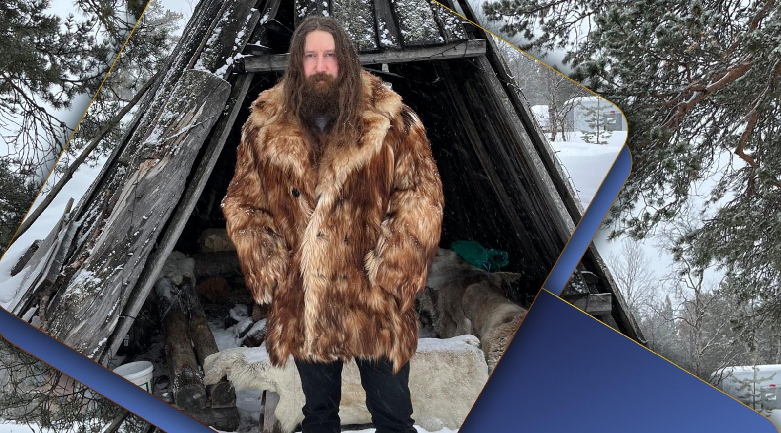 Person wearing a thick fur coat standing in front of a traditional wooden shelter in a snowy forest environment.