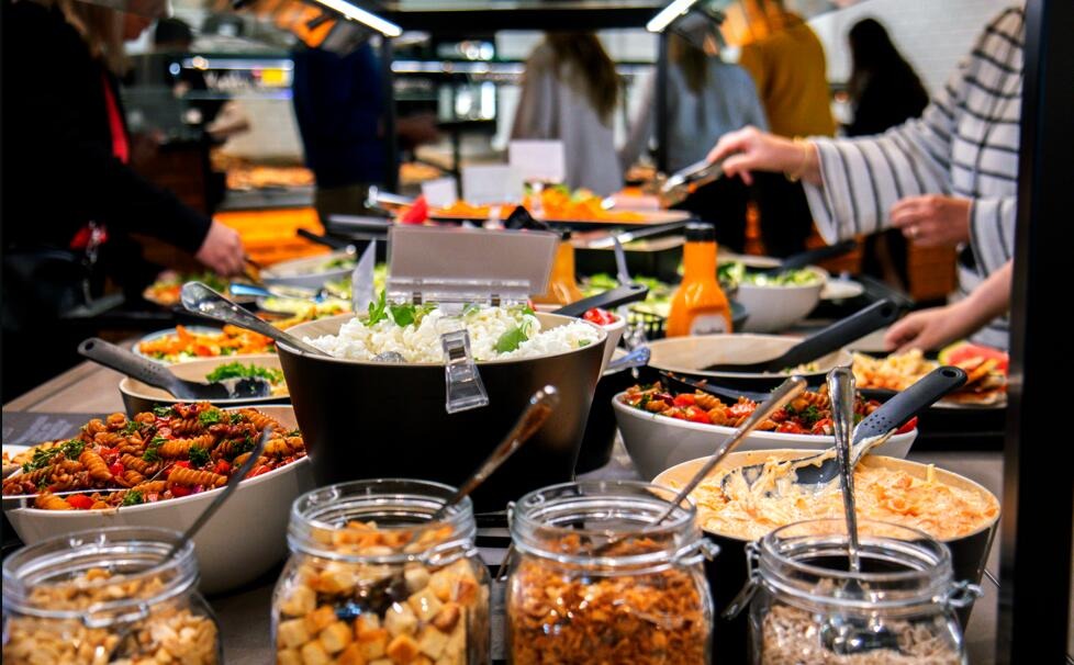 Buffébord med flera maträtter och serveringsredskap, personer som tar mat.