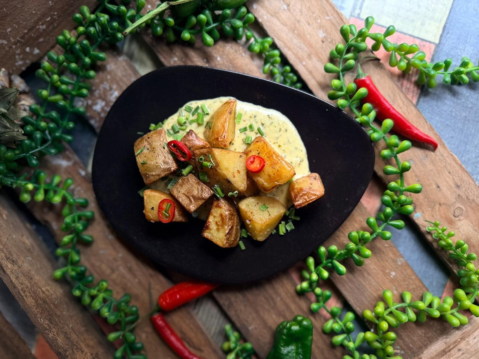 Black plate with roasted potatoes and creamy sauce, placed on wooden board.