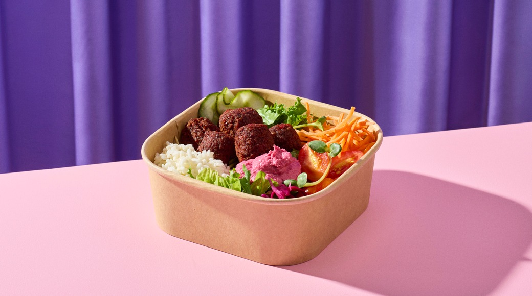 Kartonkirasiassa falafelpalloja, punajuurihummusta, riisiä, salaattia, kurkkua, tomaattia ja porkkanaraastetta. Rasia on vaaleanpunaisella pöydällä violetin verhon edessä.