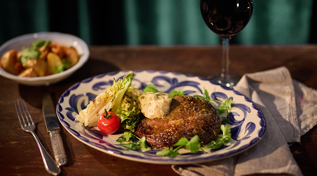 Eine, mis sisaldab lihapihvi, röstitud apteegitilli ja tomatit sini-valge mustriga taldrikul, taustaks veinipokaal ja kartulitega kauss.