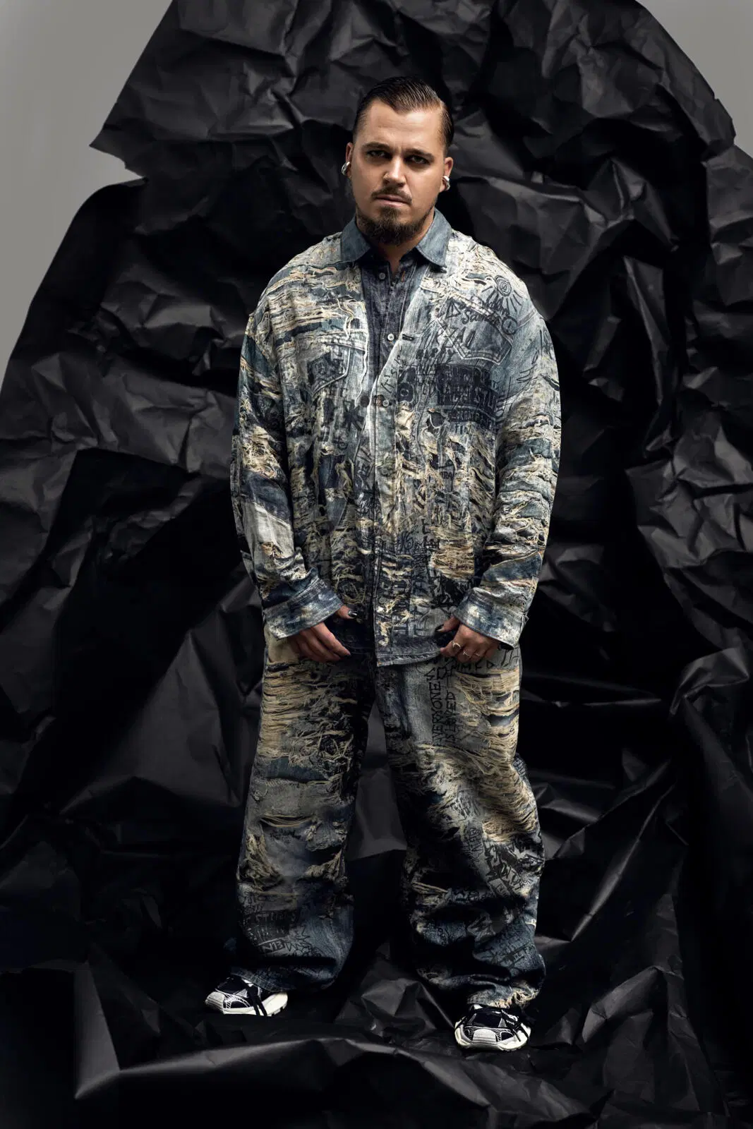 Jere Pöyhönen seisoo mustan rypystetyn taustakankaan edessä farkkuasussa ja katsoo vakavana kameraan.