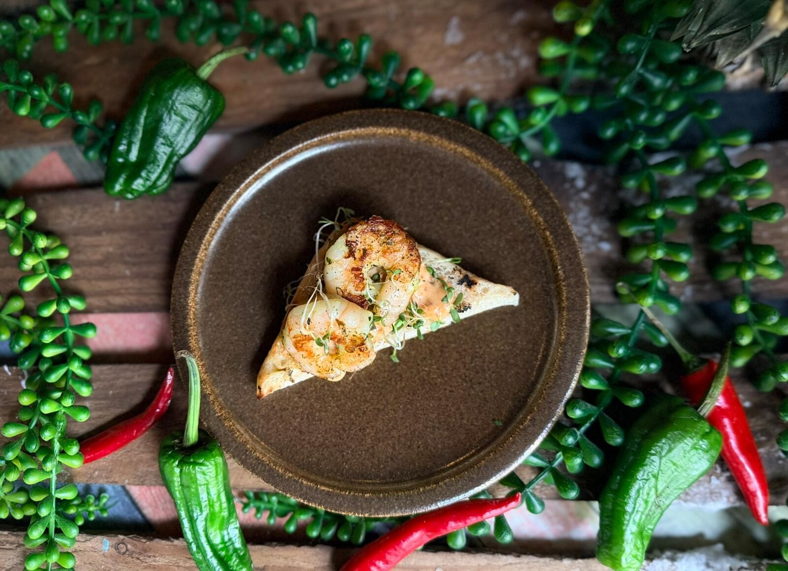 Bowl of seared prawns with roasted cauliflower, fruit and herbs on a plate. Surrounded by fresh green and red chili peppers on a rustic wooden table.