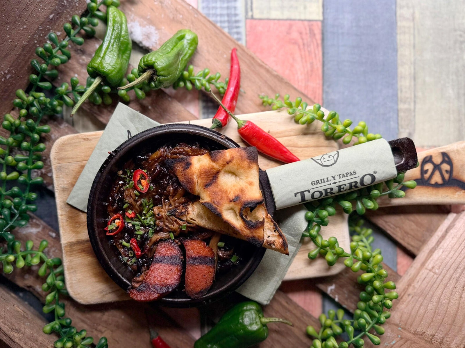 Cast iron skillet with grilled tapas featuring steak, onions, and toasted bread. Surrounded by decorative peppers and green plants.