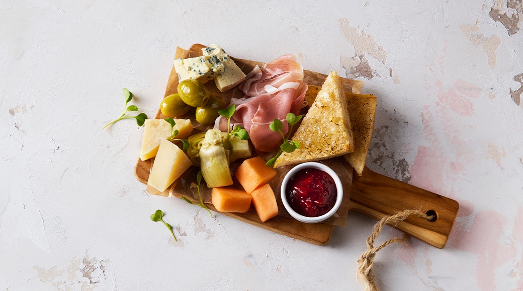 Small wooden serving board with ham, cheeses, olives, artichoke, melon, toasted bread and a small bowl of red jam.