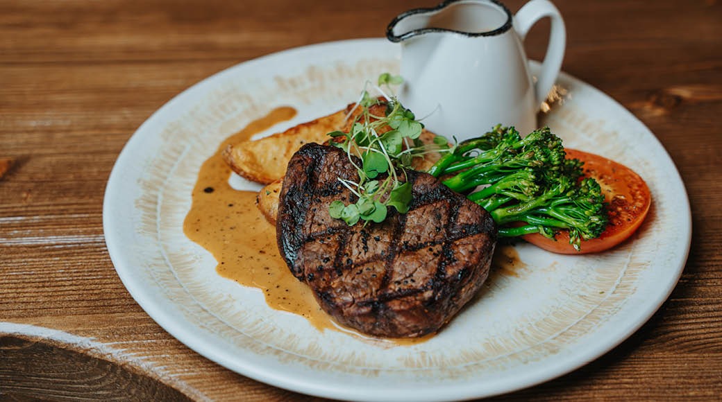 Pihviannos valkoisella lautasella, jossa grillattua lihaa, parsakaalia, tomaattia ja pieni kannullinen kastiketta puisella pöydällä.