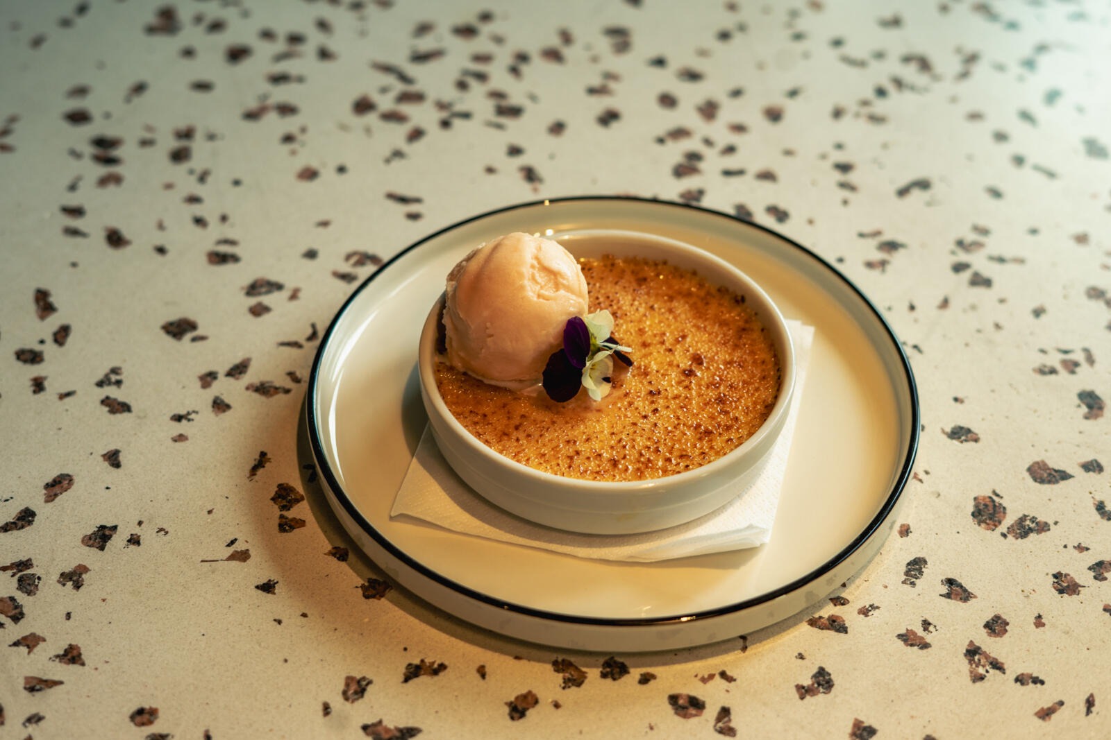 Crème brûlée lautasella, päällä pallo sorbettia ja syötävä kukka, tarjoiltuna terrasson tyylisellä pöydällä.