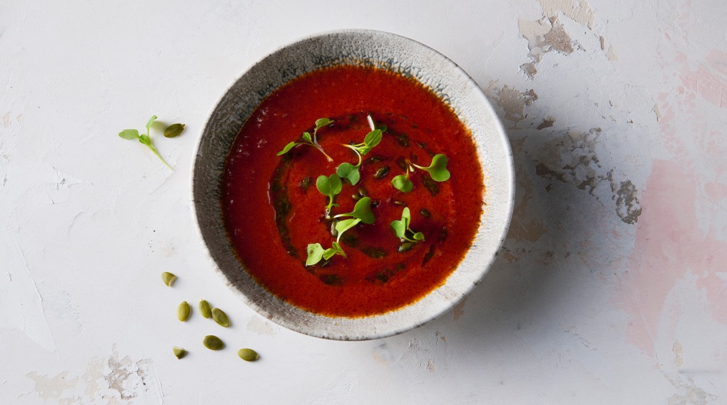 Red tomato soup in a grey bowl, topped with green microgreens and herb oil. Pumpkin seeds lie on the table.