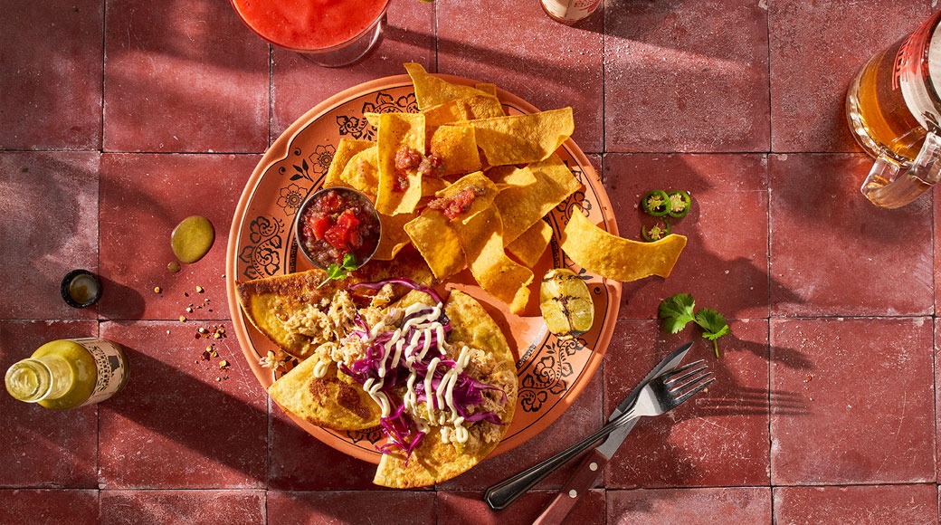 Pealtvaade kaunistatud taldrikule, millel on tortillakrõpsud, salsa ja taco punase kapsa ning kreemja kastmega. Ümber on joogid, vürtsikas kaste ja söögiriistad punasel plaadil.