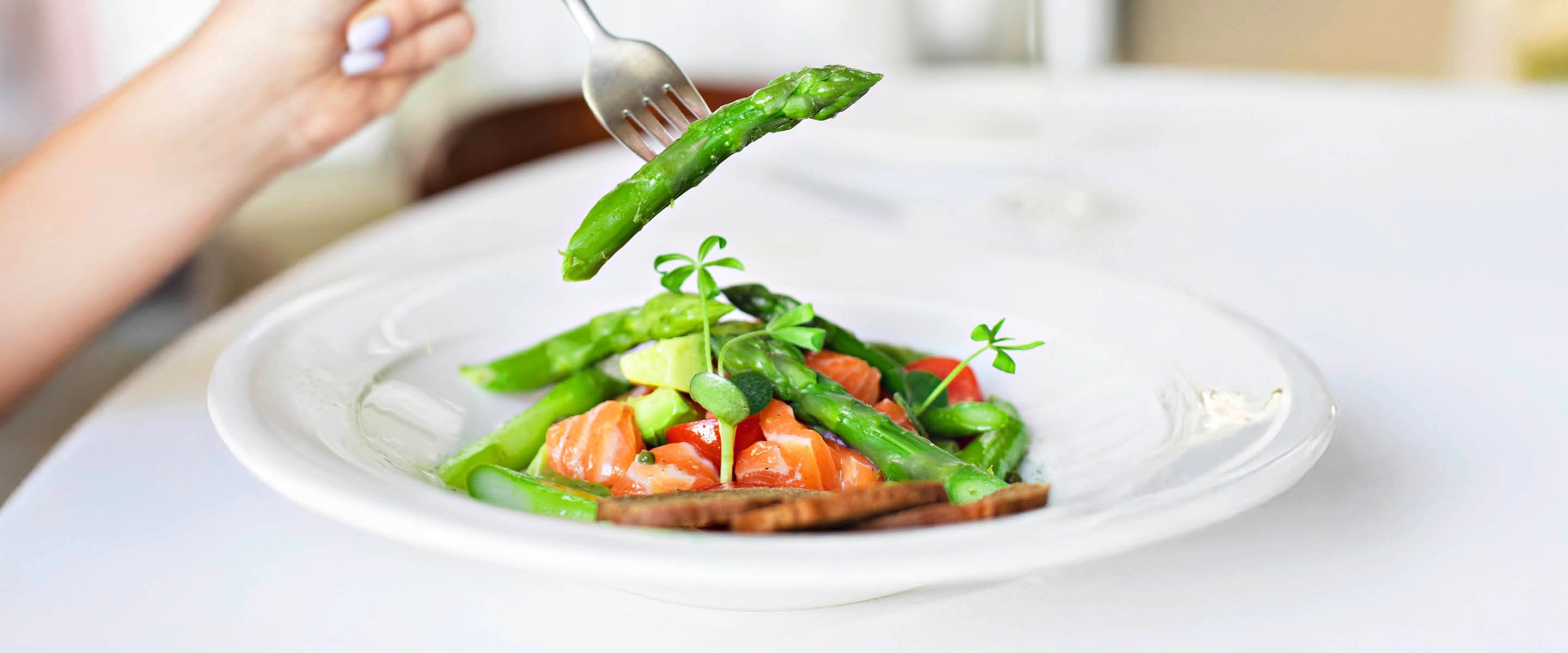 Green asparagus on a fork above a white plate containing salad with salmon, asparagus and microgreens.