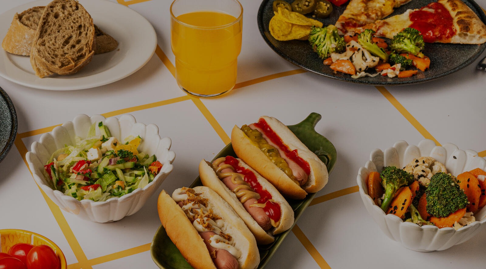 Diverse food spread featuring hot dogs, salads, vegetables, pizza, bread and orange juice on a white table with yellow lines.