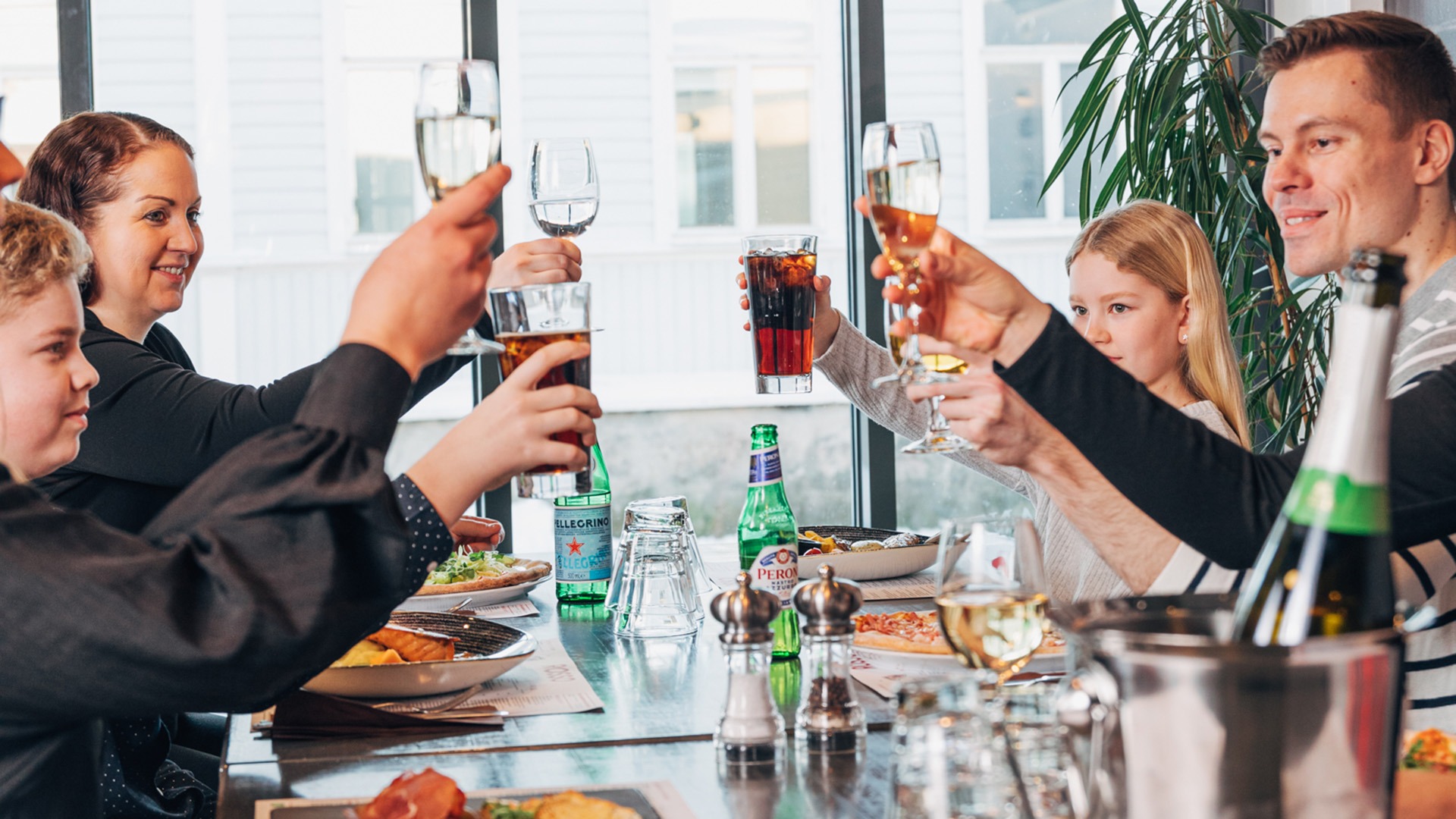 Familj skålar tillsammans på en restaurang. På bordet finns mat, drycker och vattenflaskor. Stora fönster och en grön växt syns i bakgrunden.
