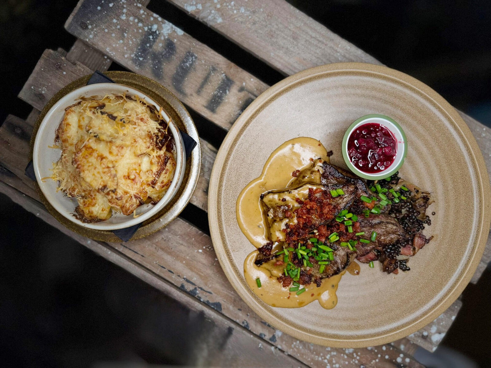 Two dishes: meat main course with sauce, chopped chives and lingonberry jam, accompanied by a cheesy gratin side dish.