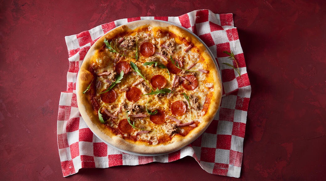 Pizza topped with pepperoni, ham, tuna, cheese and rocket. The pizza sits on a red and white checkered cloth on a red table.