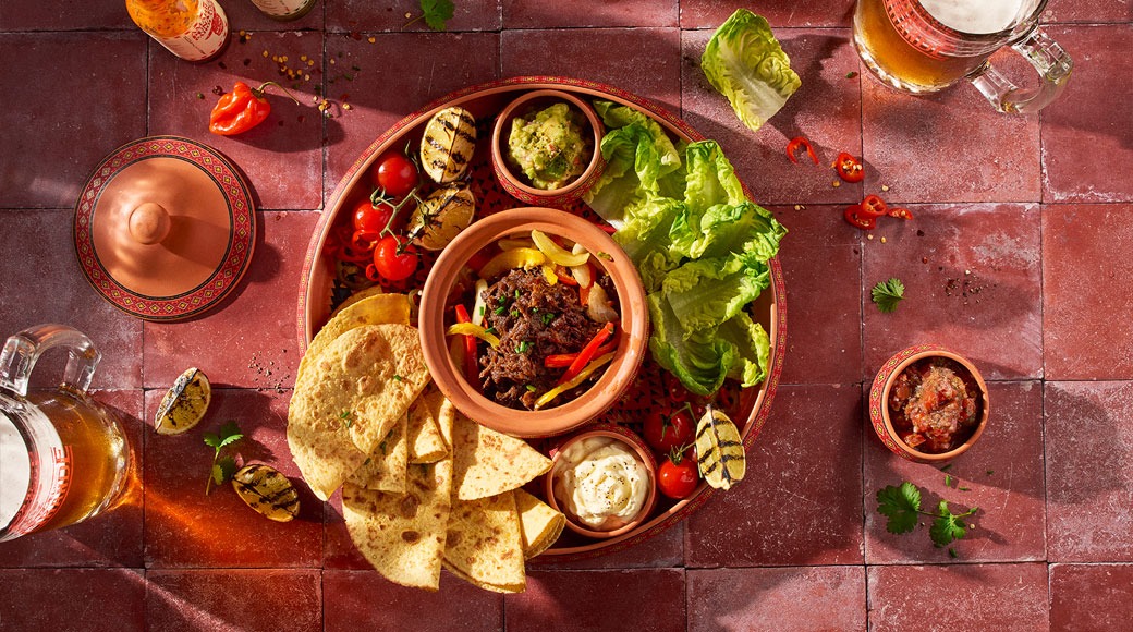 Mehhiko road ümaral serveerimisalusel: kitsas liha paprikatega, tortillakolmnurgad, salat, guacamole, hapukoor ja salsa. Kõrval on õlleklaasid.
