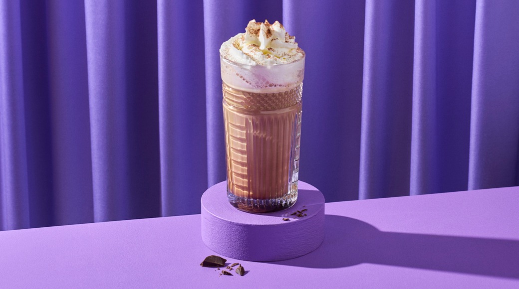 Kuviollinen lasi kuumaa suklaata, päällä kermavaahtoa ja kaakaojauhetta. Lasi on violetilla jalustalla violettia verhoa vasten.
