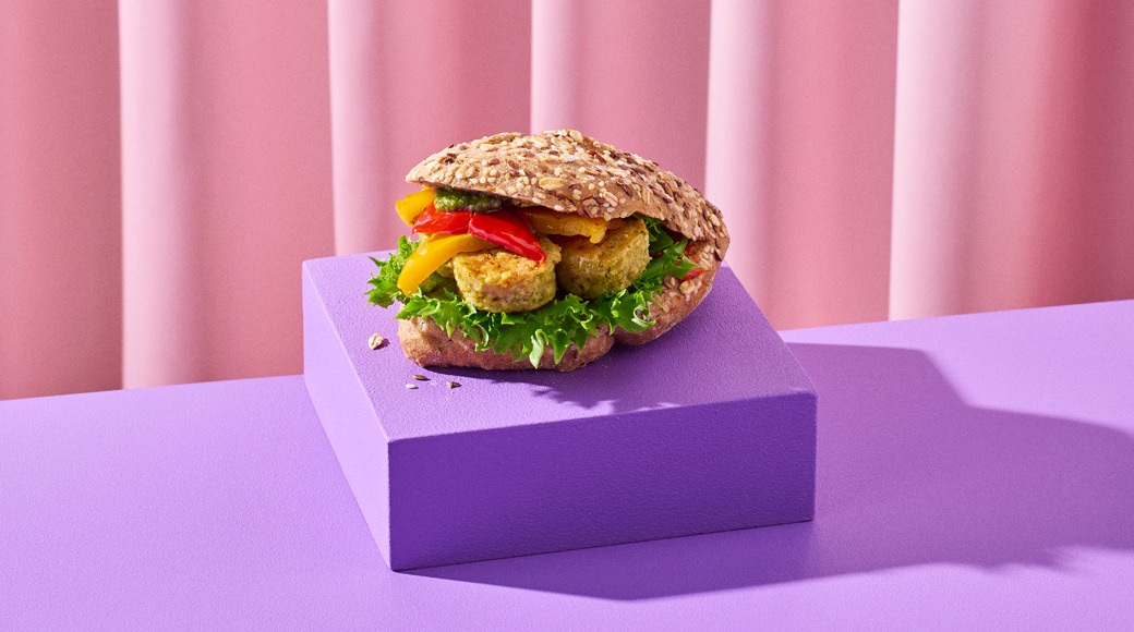 Täysjyväsämpylä, jonka välissä on falafelpihvejä, salaattia ja paprikaa, violetilla korokkeella vaaleanpunaisen verhon edessä.