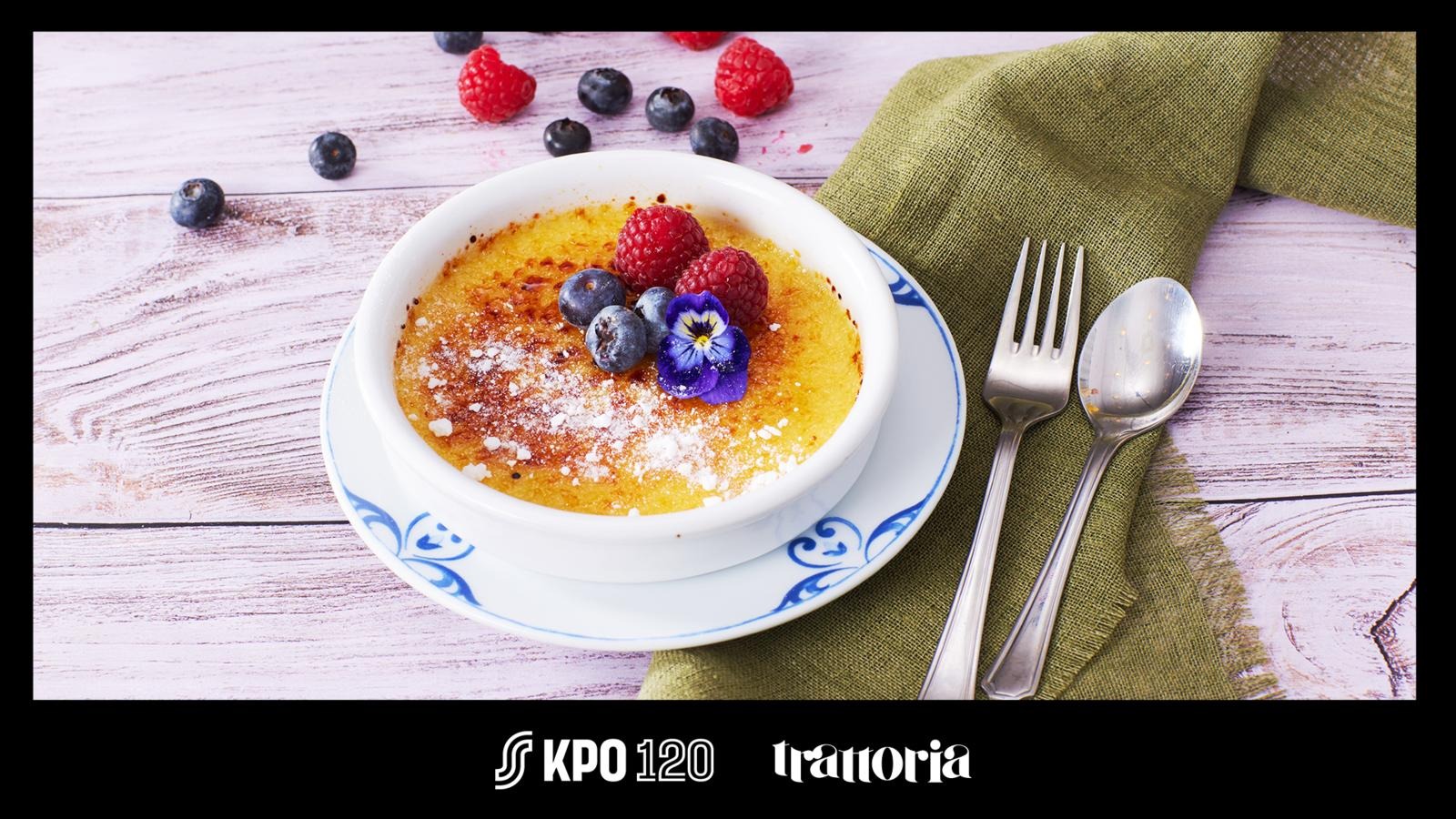 Crème brûlée garnished with blueberries, raspberries and an edible flower. Fork, spoon and green napkin beside it on white wooden surface.