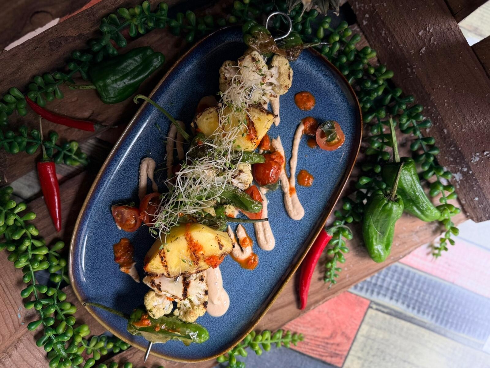 Grilled vegetable skewers on a blue plate with potato, cauliflower florets and peppers, garnished with sprouts and sauce.