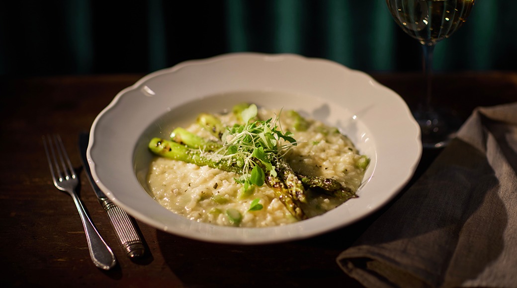 Valge taldrik spargli risotoga, mille peal on spargli võrsed ja mikrorohelised. Kõrval kahvel ja veinipokaal tumeda laua peal.