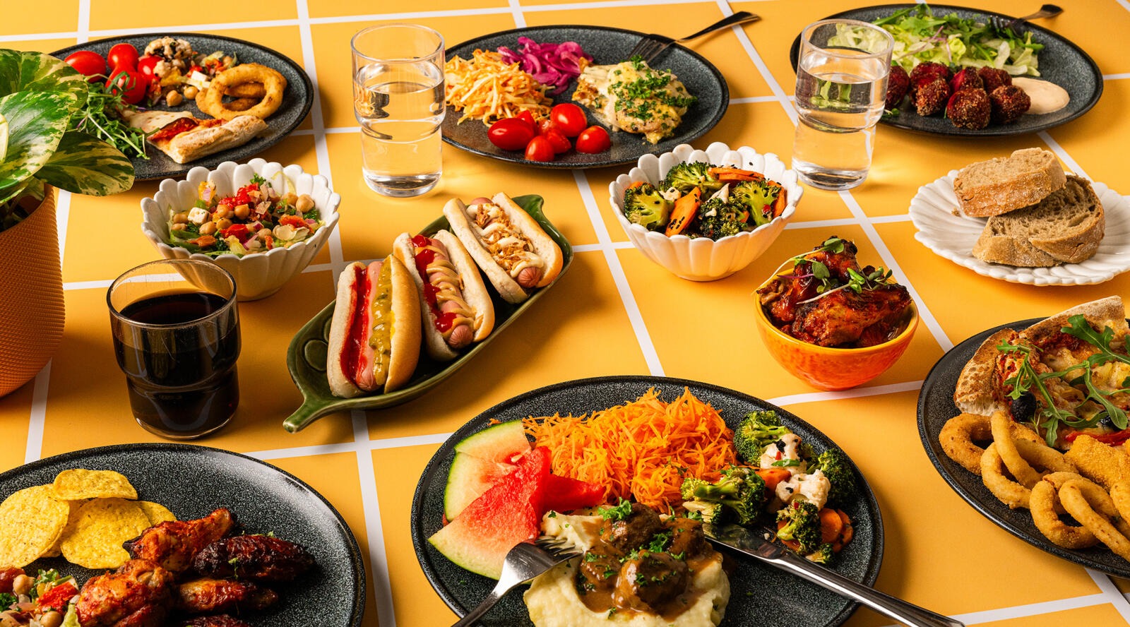 Monipuolinen ruokapöytä, jossa on hotdogeja, salaatteja, sipulirenkaita, meatballeja, perunasosetta, kasviksia ja leipää keltaisella pöydällä vesijuomien kera.