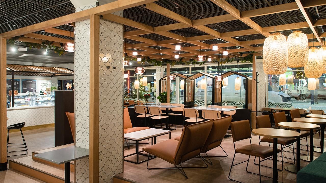Coffee Housen avara sisätila, jossa puuelementit, vihreät kasvit ja roikkuvat valaisimet luovat lämpimän ja kutsuvan tunnelman.