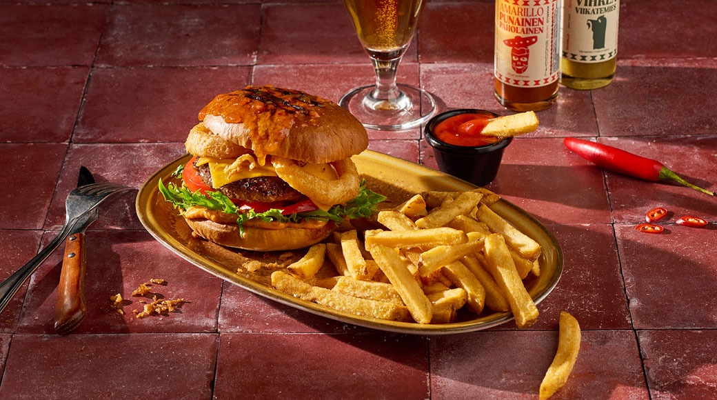 Burger juustu, salati, tomati ja sibularõngaga ning friikartulid ovaalsel taldrikul. Kõrval on dipikauss, õlleklaas, vürtsikaste pudelid ja tšillid punasel plaaditud laual.