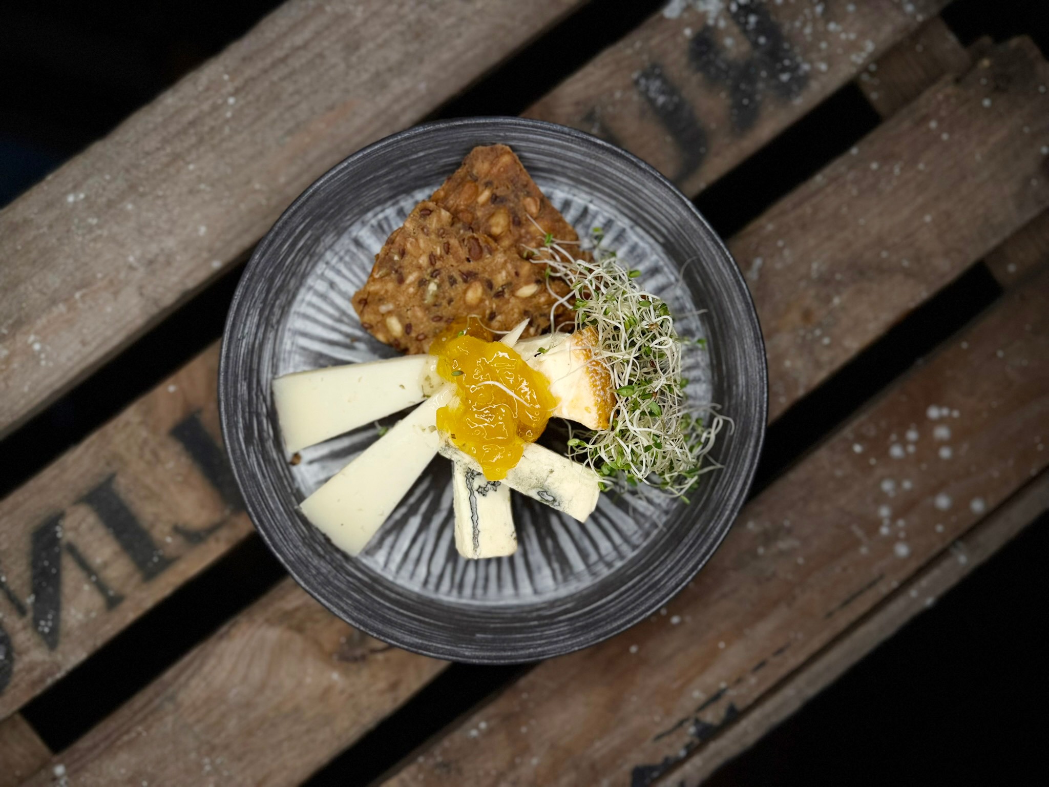 Cheese plate with seeded cracker, cheese wedges, fruit preserve, and sprouts. Served on wooden slats.
