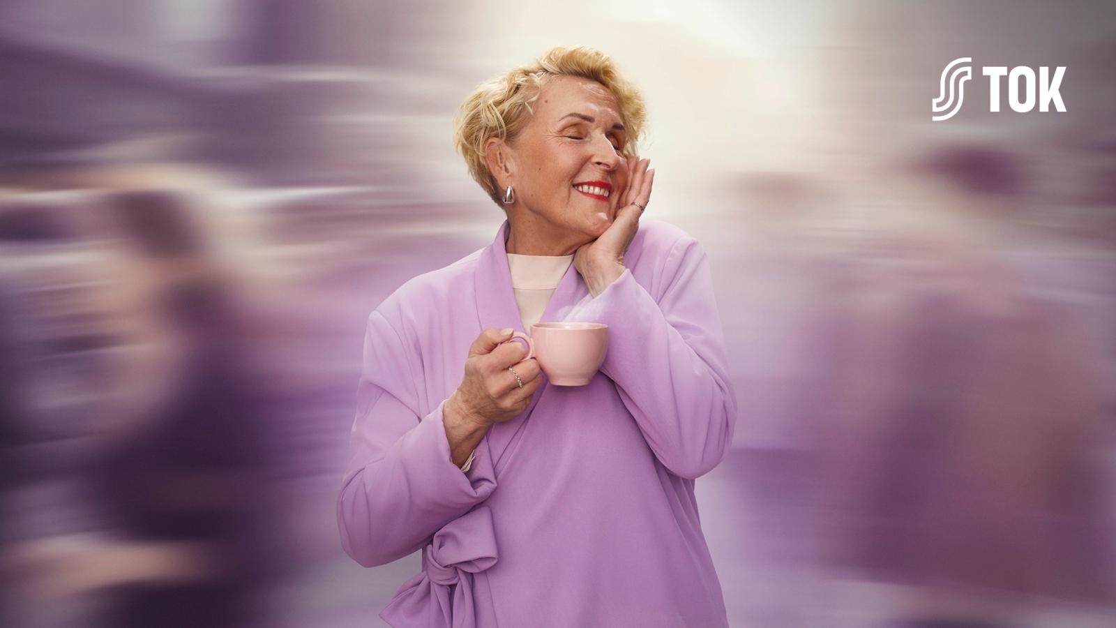 Äldre person i ljusrosa dräkt ler medan hen håller en rosa kaffekopp. Bakgrunden är suddig i lila toner.
