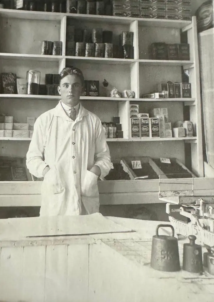 Vanha mustavalkokuva, jossa henkilö seisoo valkoisessa takissa myymälässä.