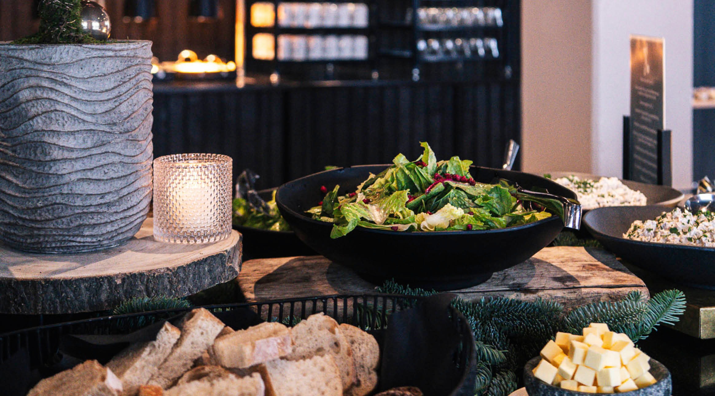 Buffet spread featuring green salad in a black bowl, cheese cubes and bread. Wooden serving board decorated with candles and evergreen branches.