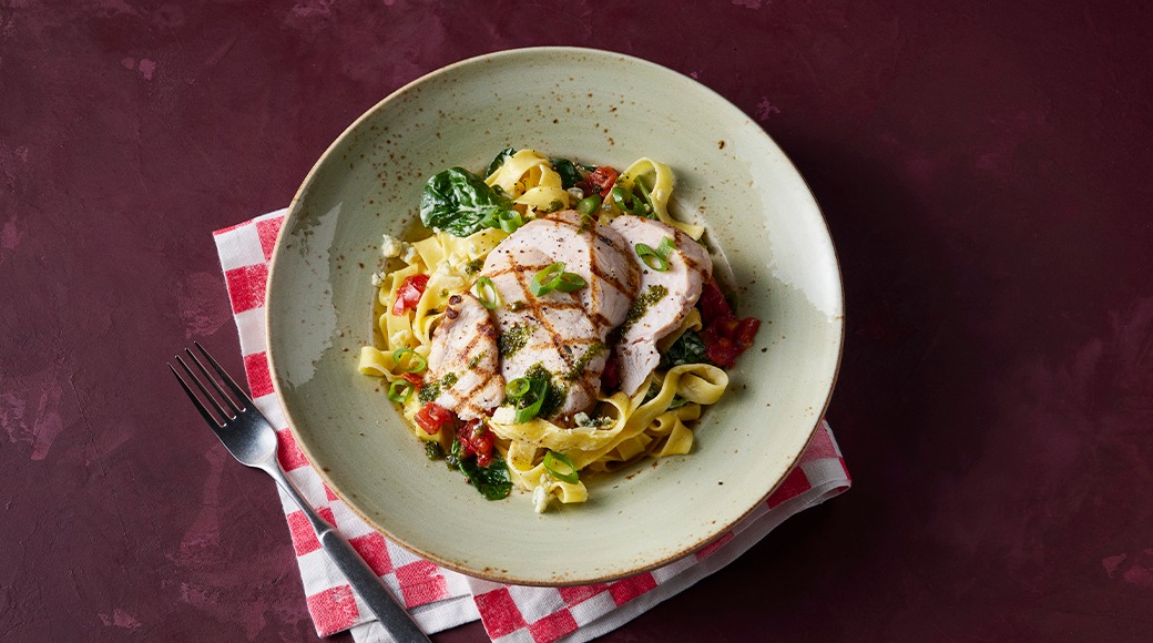 Bowl of tagliatelle with grilled chicken, spinach, tomatoes and spring onion. The bowl sits on a red checked napkin next to a fork.