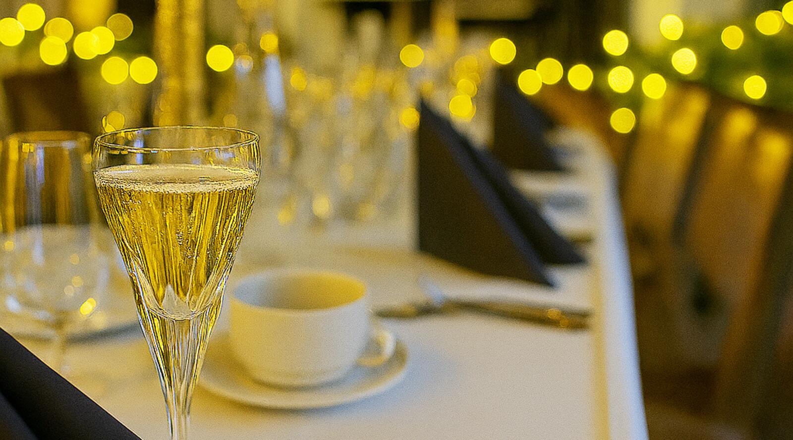 Crystal champagne glass on table, with place settings and golden lights in background.