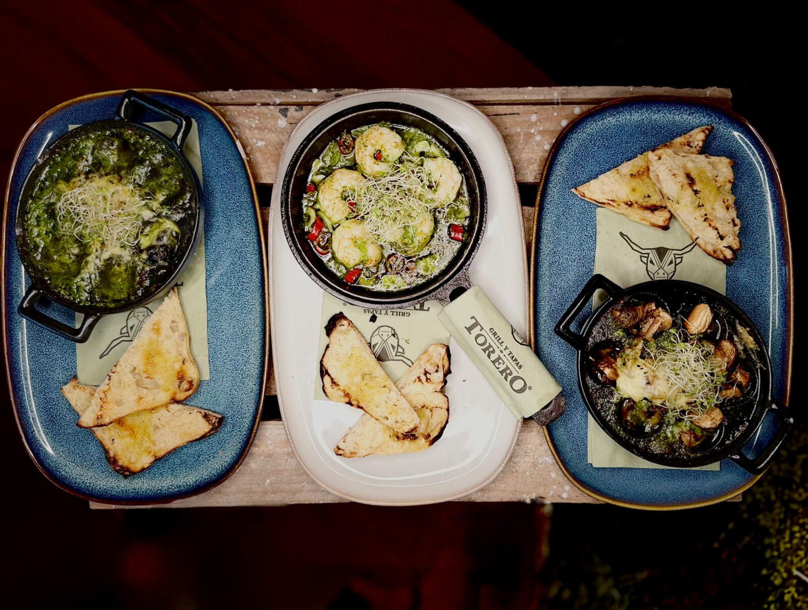 Tray of tapas featuring three small pans with different dishes and four triangles of bread on blue and white plates.