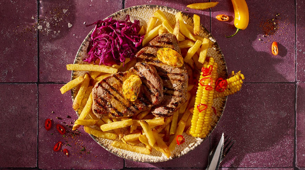 Grillattua pihviä, jonka päällä on maustevoita, sekä ranskalaisia, punakaalisalaattia ja maissia lautasella. Chilisilppua ja keltainen chili purppuranpunaisella laatalla.