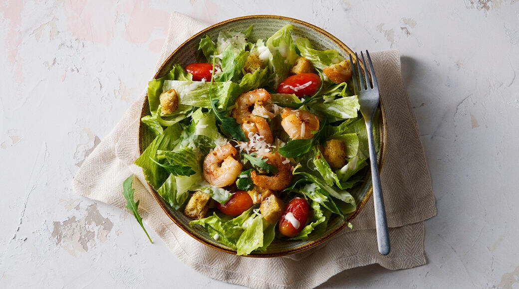 Bowl of leafy salad with shrimp, cherry tomatoes, croutons and grated cheese. A light cloth lies under the bowl, with a fork beside it.
