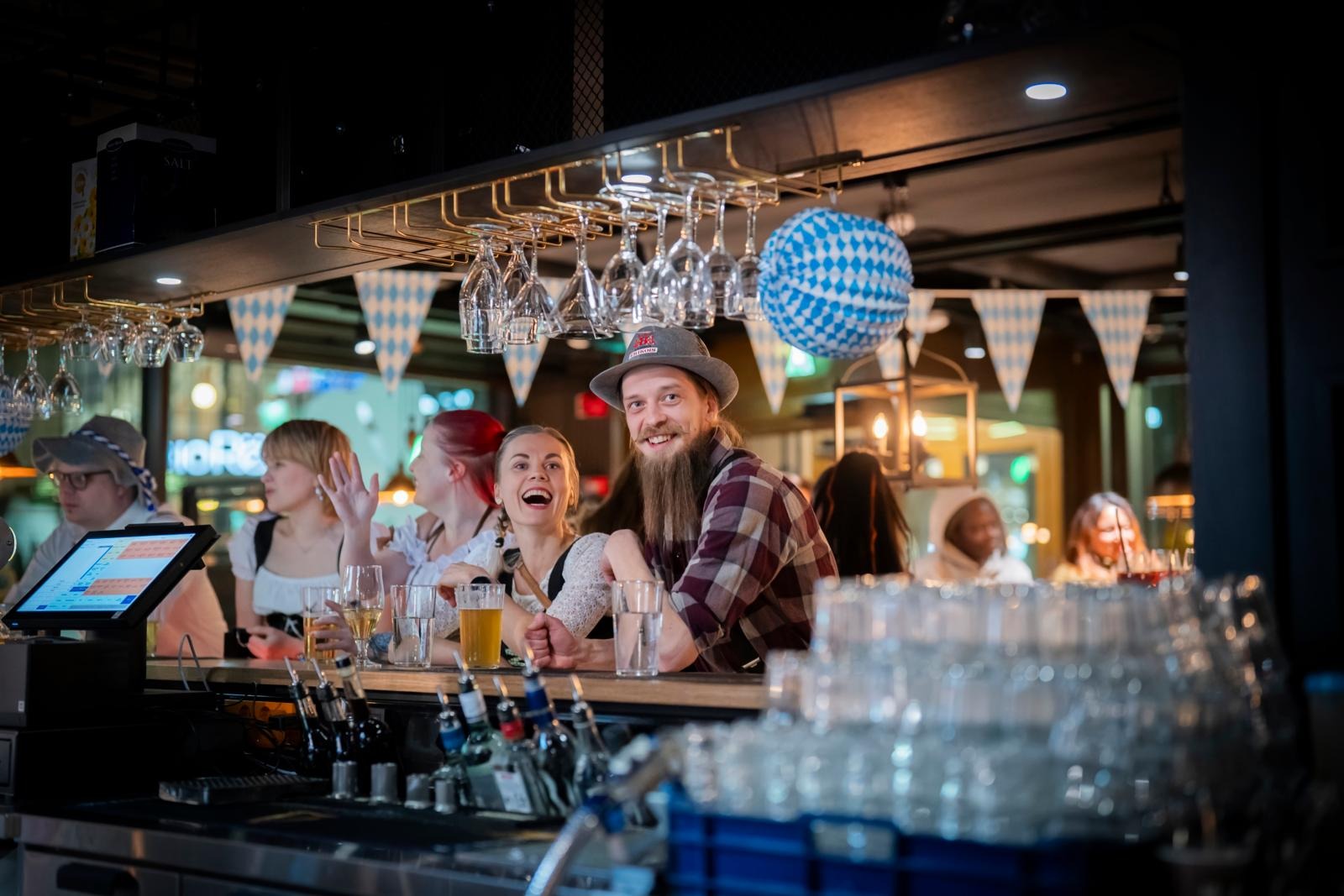 Baari, jossa ihmiset nauttivat juomia. Sinivalkoiset Oktoberfest-koristeet, laseja roikkuu ylhäällä baaritiskin yläpuolella.