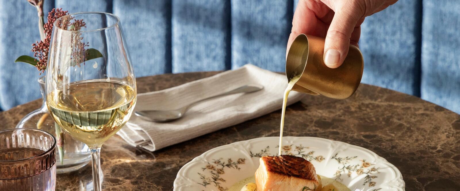 Käsi kaataa pienestä messinkikannusta kastiketta kauniisti asetellun kala-annoksen päälle. Pöydällä on viinilasi ja servetti.