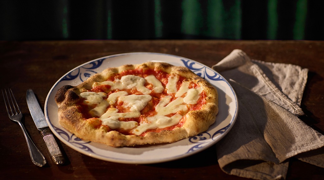 Margheritapizza sinikoristekuvioisella lautasella puisella pöydällä, vieressä haarukka, veitsi ja pellavaservetti.