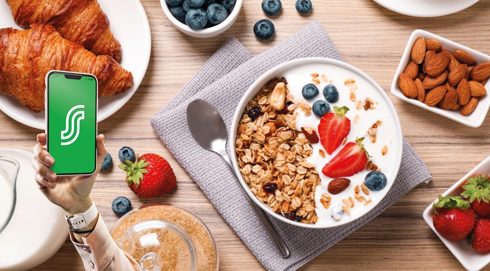 Hommikusöök jogurti, müsli, marjade, mandlite ja croissant'idega. Käsi hoiab nutitelefoni, millel on roheline rakendus.