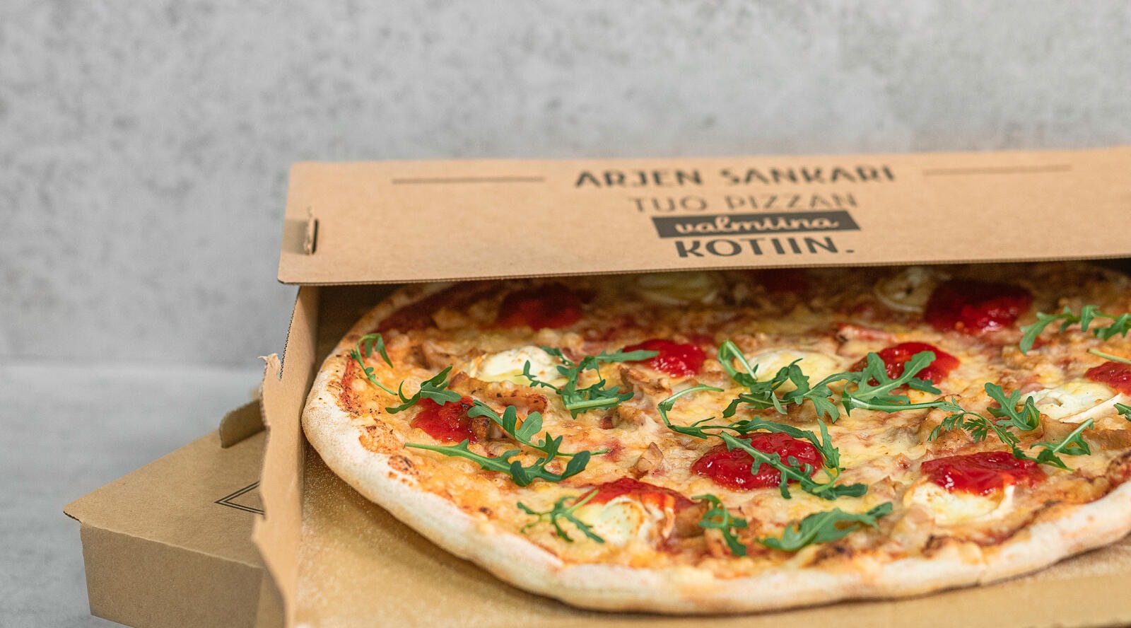 Pizza laatikossa, päällä tomaattia, juustoa ja tuoreita rucolanlehtiä.