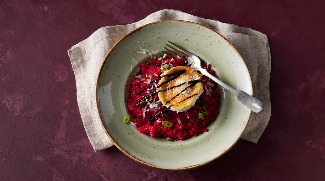 Beetroot risotto in a green bowl, topped with grilled cheese, balsamic glaze, spring onion and grated cheese.