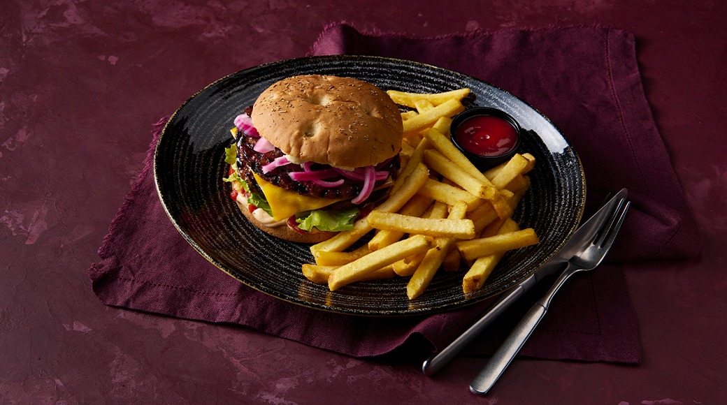 Burger with cheese, lettuce and red onion, served with fries and ketchup on a black plate. A knife and fork lie beside it.