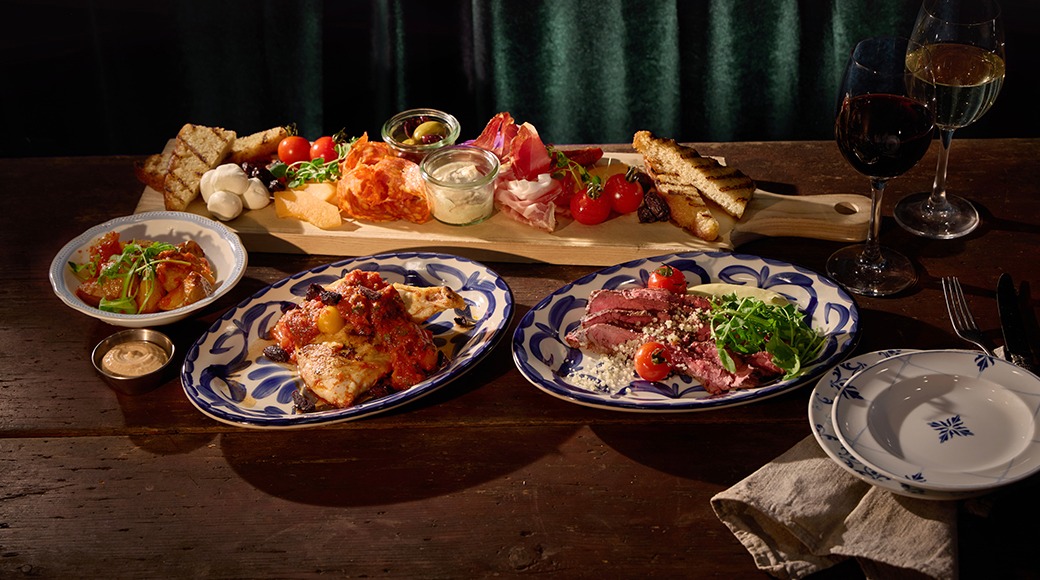 Laud rikkaliku toiduga: taldrikutel liha ja kala, lõikelaual juustud, lihalõigud ja leib. Kaks veiniklaasi täiendavad kaetud lauda.