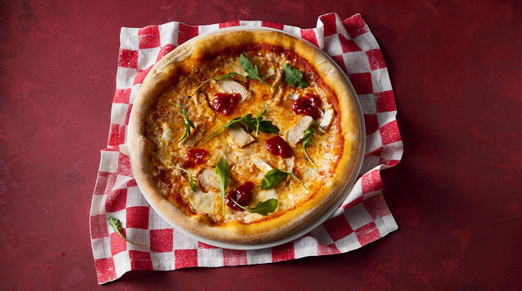 Pizza topped with chicken, rocket and dollops of red sauce on a white plate over a red and white checkered cloth.