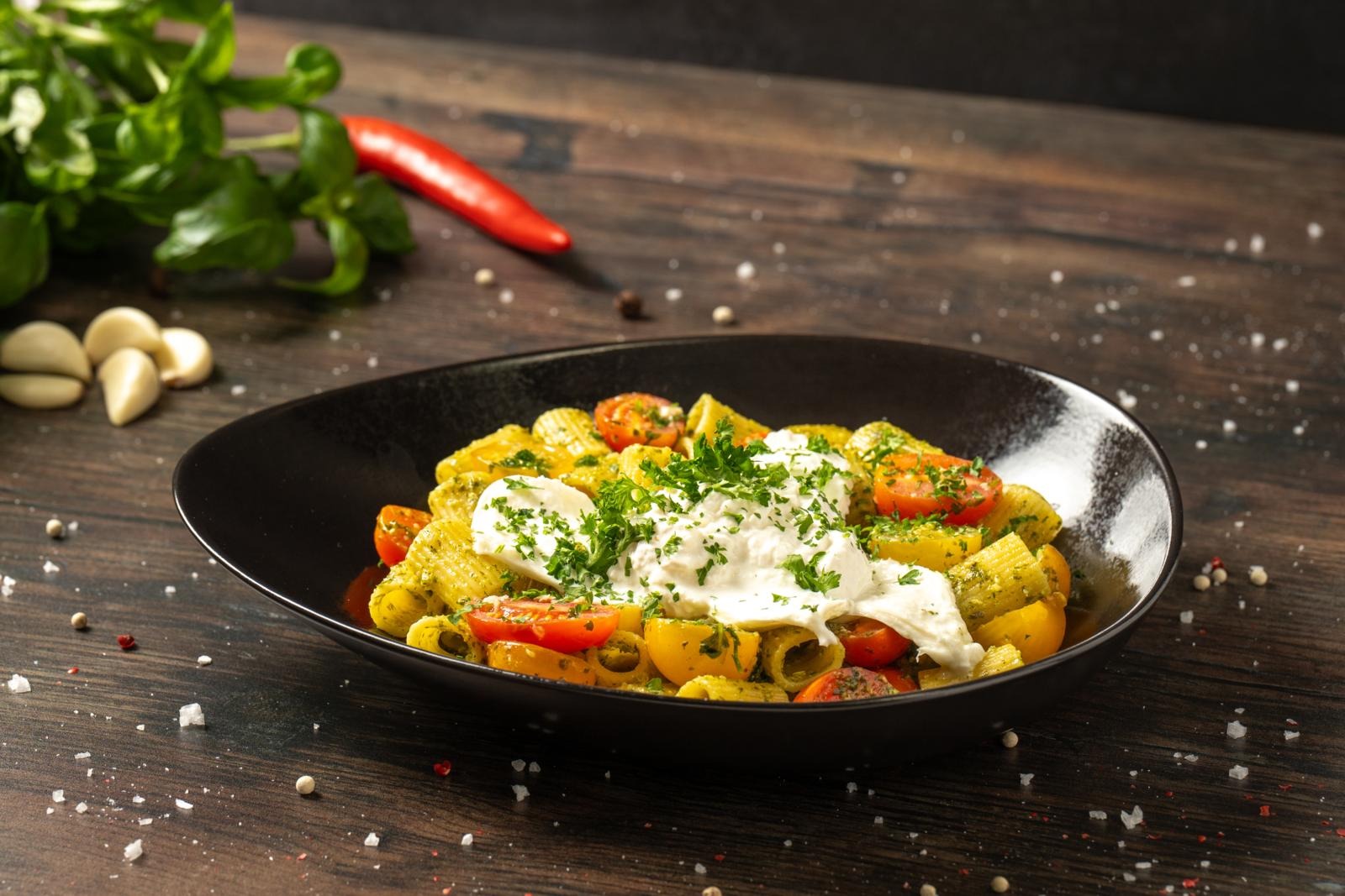 Mustal taldrikul pasta, kirsiktomatid ja mozzarella ürdikastmes. Taustal värske basiilik, küüslauguküüned ja punane tšilli.