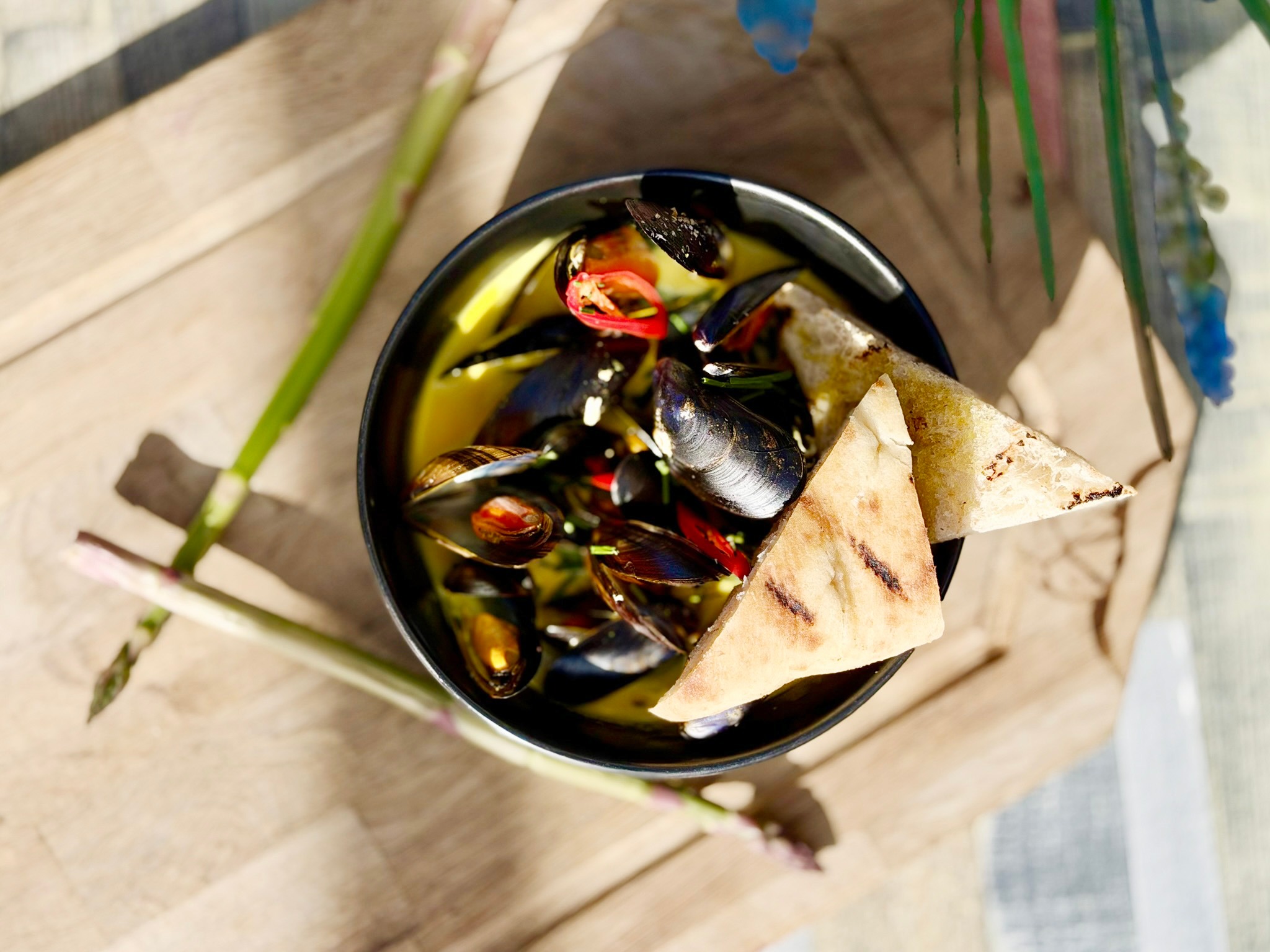 Mussel dish in black bowl with chili peppers and grilled bread triangles, served on a wooden board.